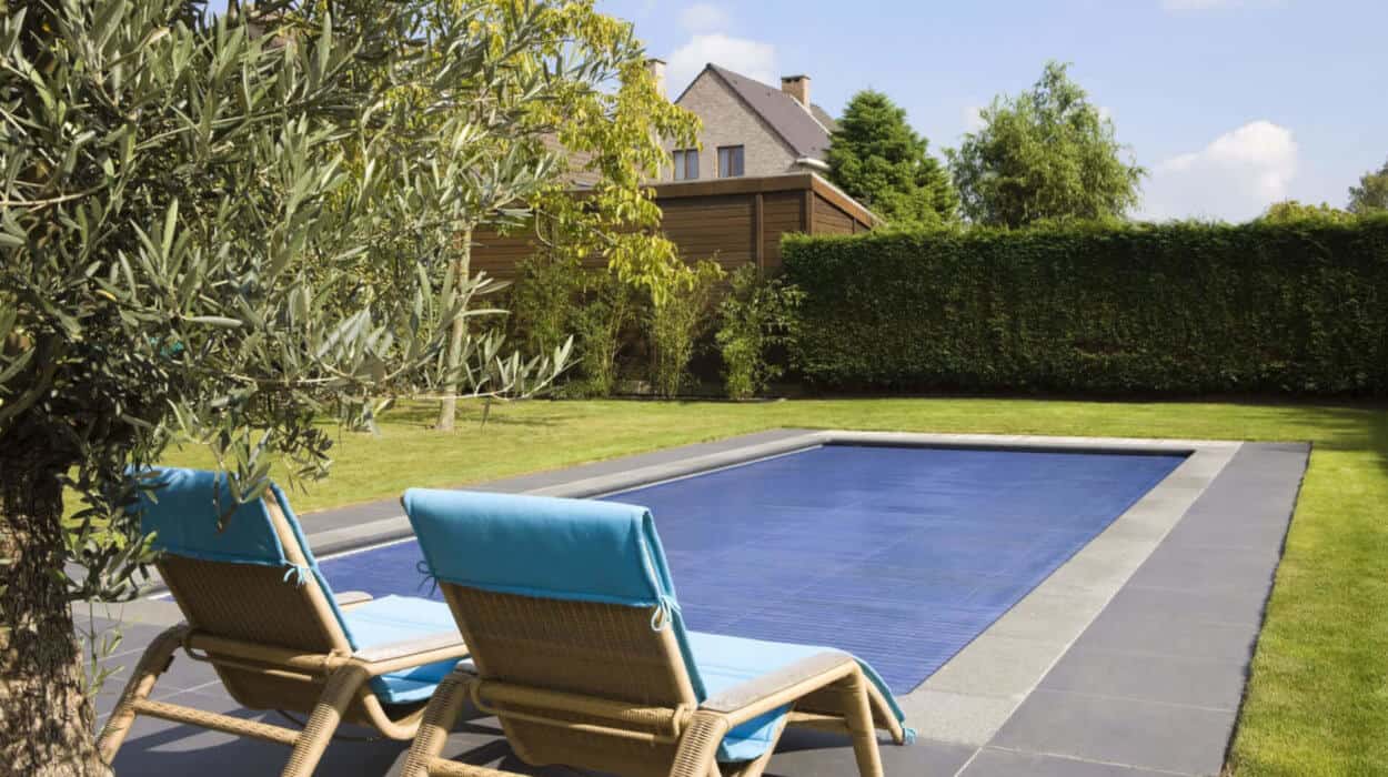 Two wicker lounge chairs with blue cushions face a covered rectangular pool, surrounded by greenery and a hedge. A house is visible in the background.