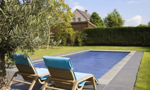 Two wicker lounge chairs with blue cushions face a covered rectangular pool, surrounded by greenery and a hedge. A house is visible in the background.