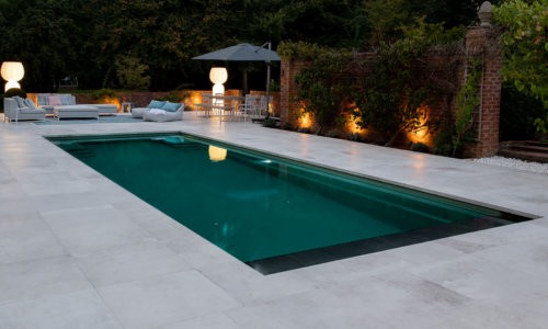 Evening view of a luxury outdoor swimming pool with soft garden lighting, modern patio furniture, and surrounding trees.