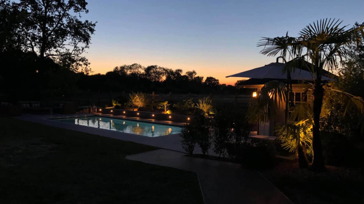 Outdoor swimming pool lit at sunset, with palm trees and garden lighting.