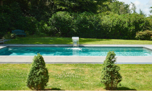 Luxury rectangular garden pool with a central waterfall feature and lush greenery in the background.